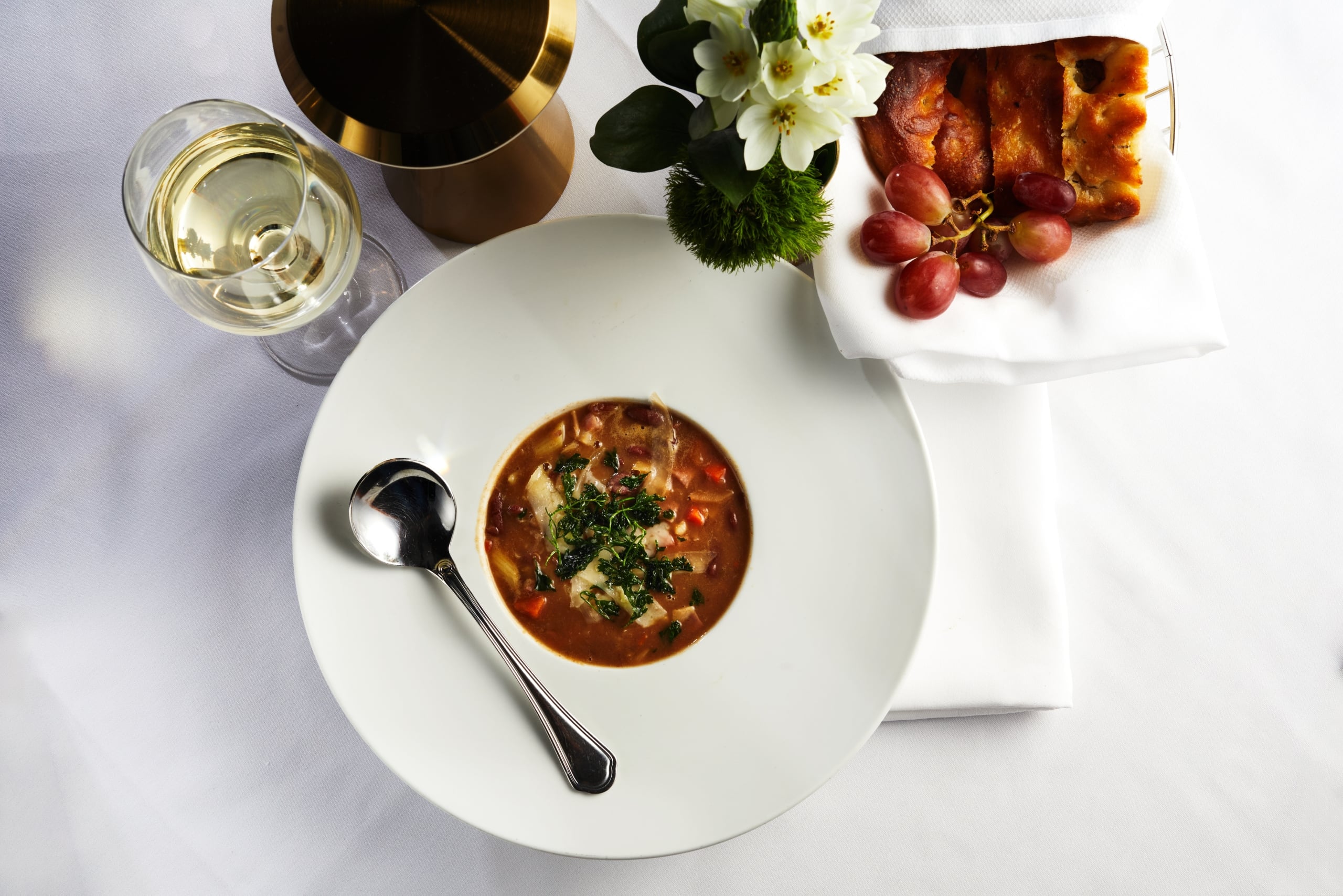 A refined meal setting with savory soup, white wine, fresh focaccia, and grapes.