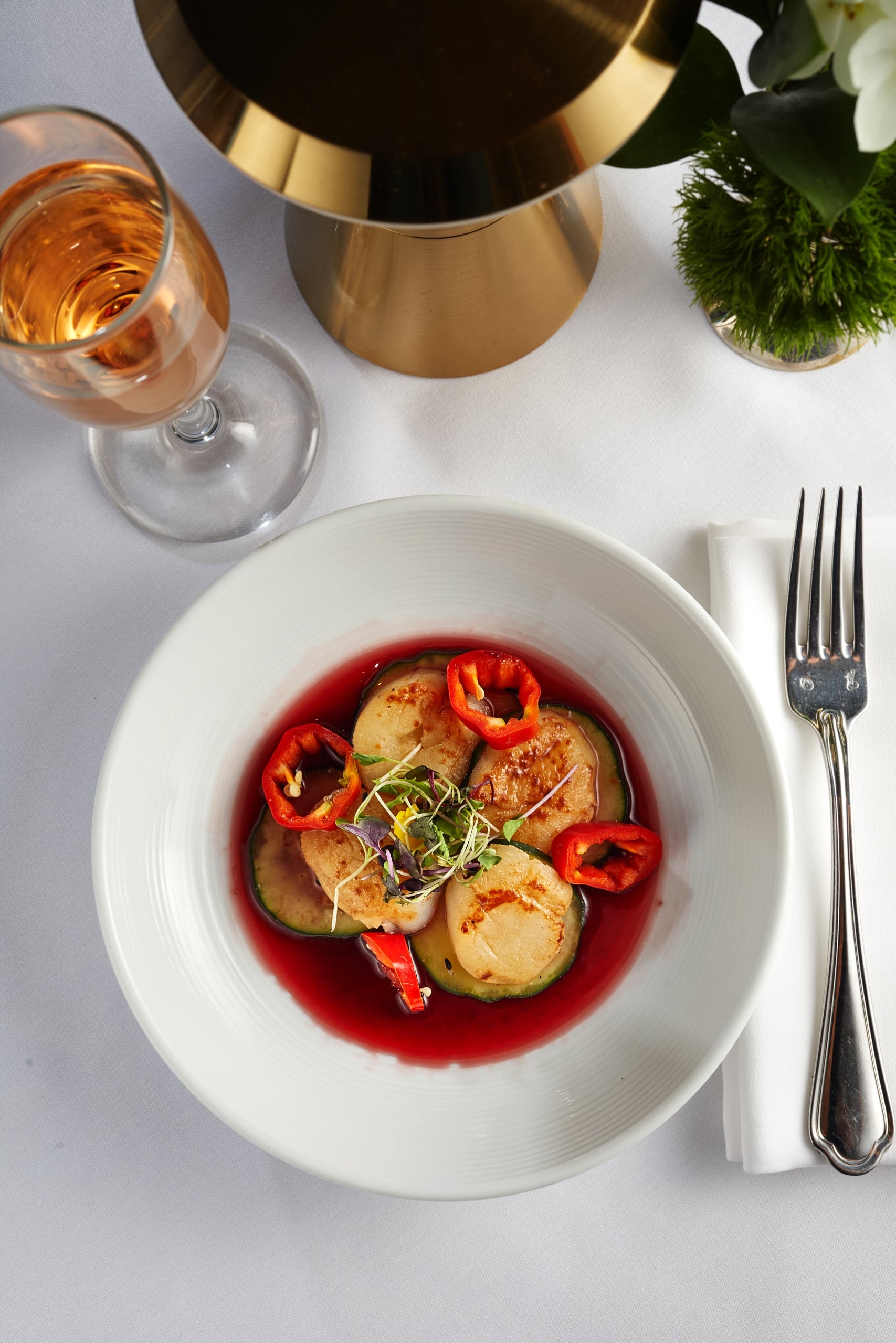 Elegant plate of seared scallops, zucchini, red peppers in rich red sauce, garnished with microgreens and rosé.