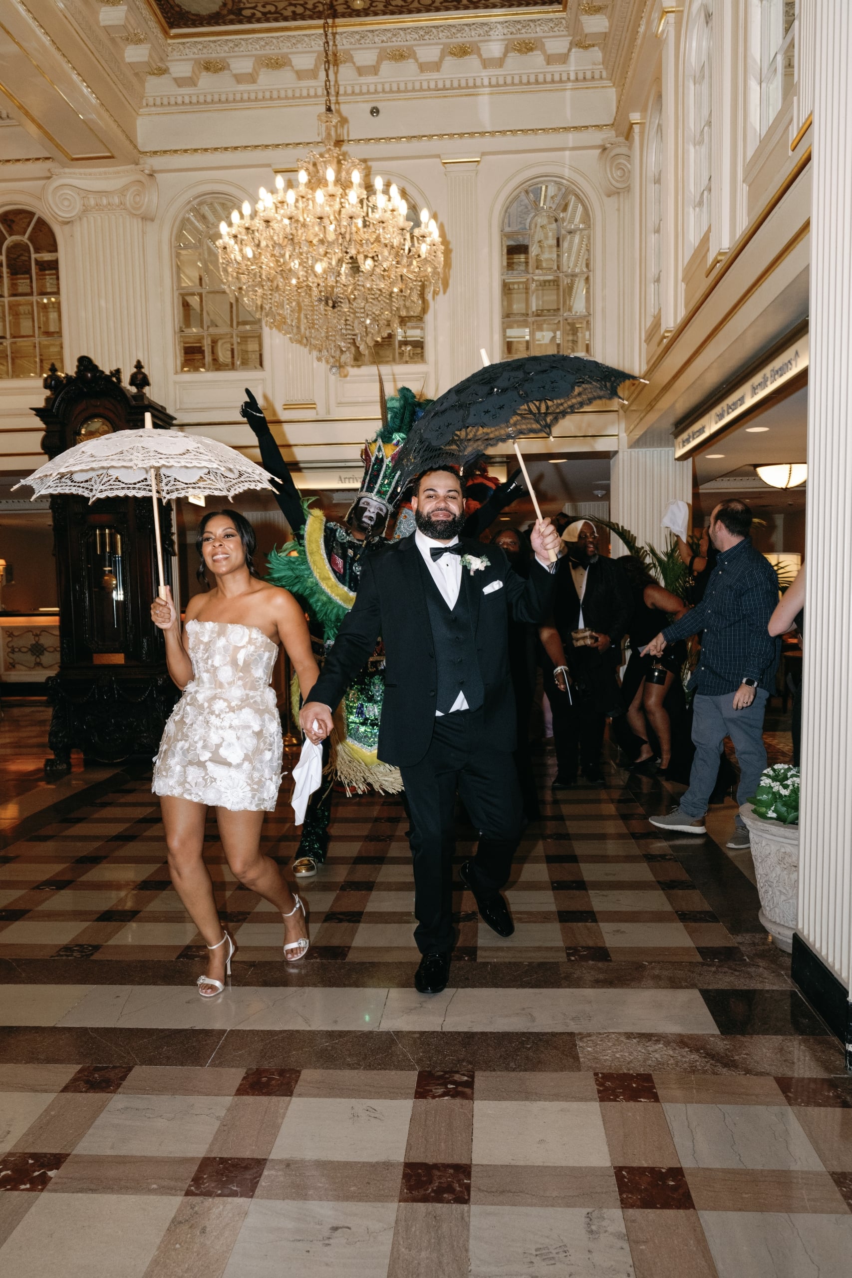 Bride and groom with decorative umbrellas lead a lively Second Line parade in a grand New Orleans hotel lobby.