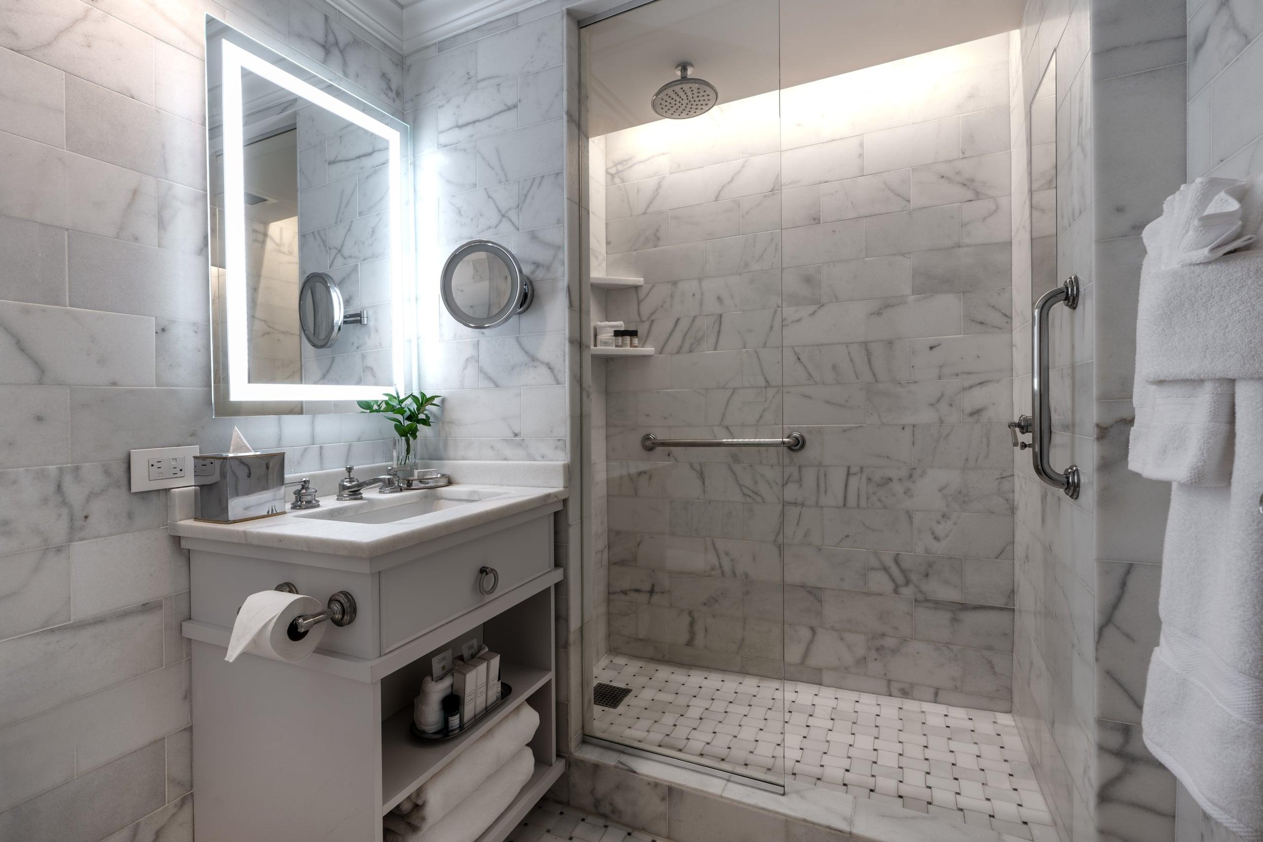 Modern marble bathroom in a New Orleans hotel, featuring a vanity, lighted mirror, and glass walk-in shower.