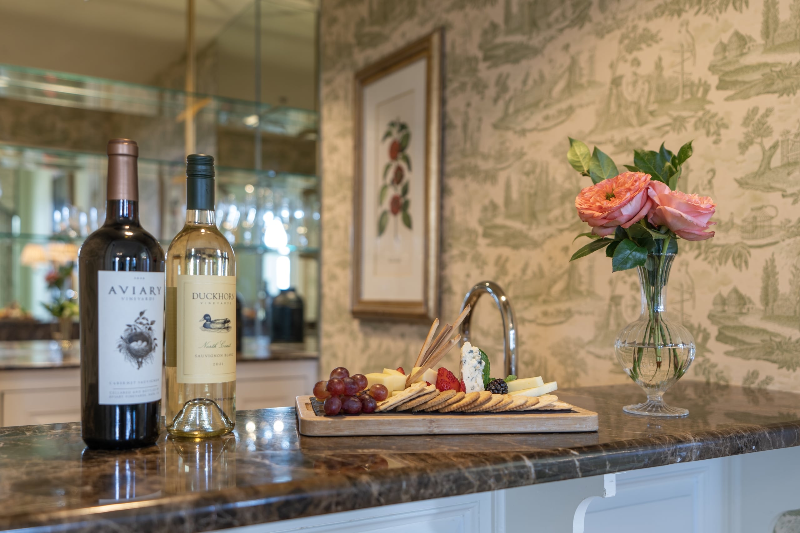 Premium red and white wines, a gourmet cheese and fruit board, and fresh pink roses on a marble counter.