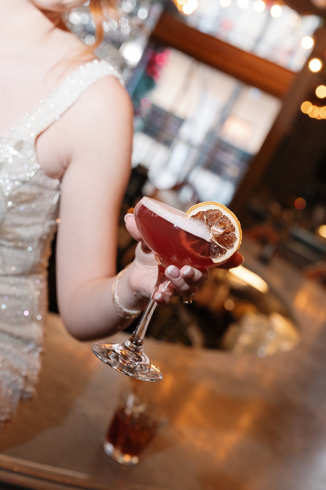 Guest in a sequined dress holding a red craft cocktail garnished with a dried orange slice at a New Orleans bar.