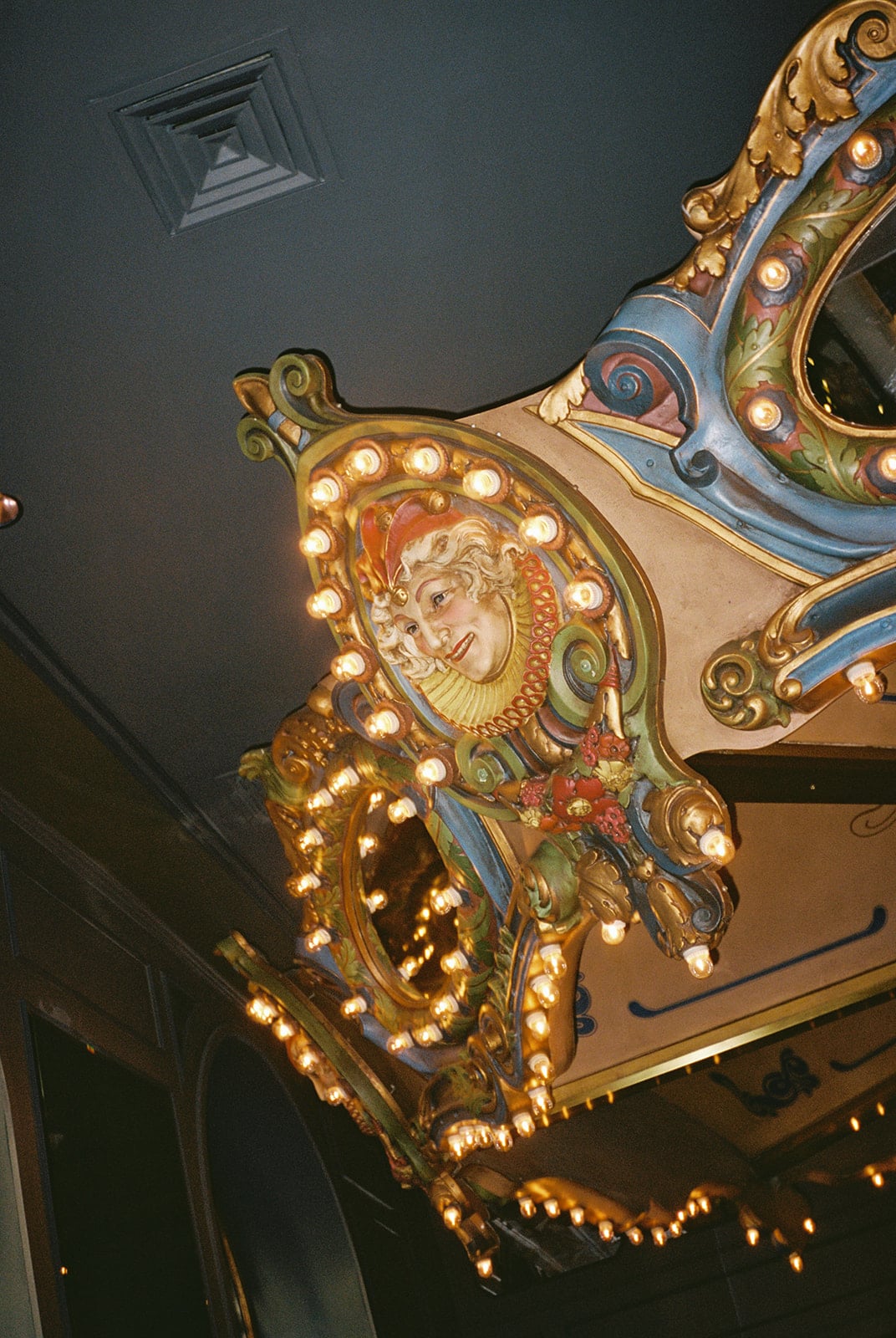 Ornate, brightly lit vintage carousel panel featuring a smiling jester and colorful decorations, a New Orleans attraction.