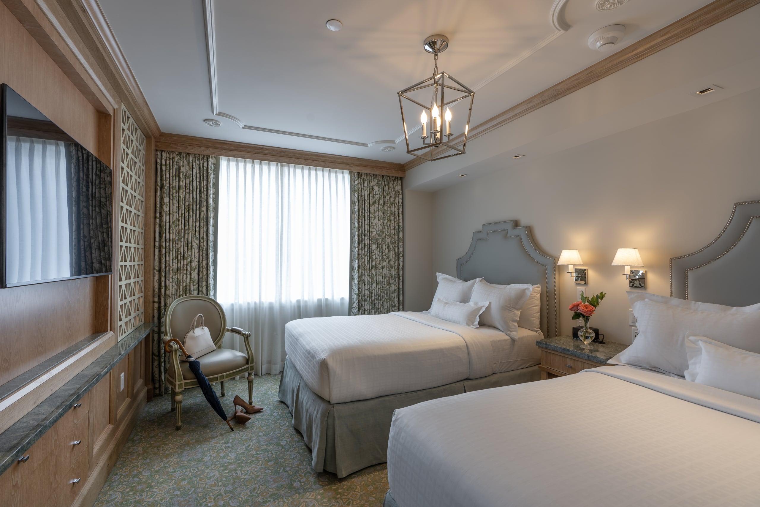 Elegant New Orleans hotel room with two plush beds, patterned carpet, bright window, and sophisticated decor.