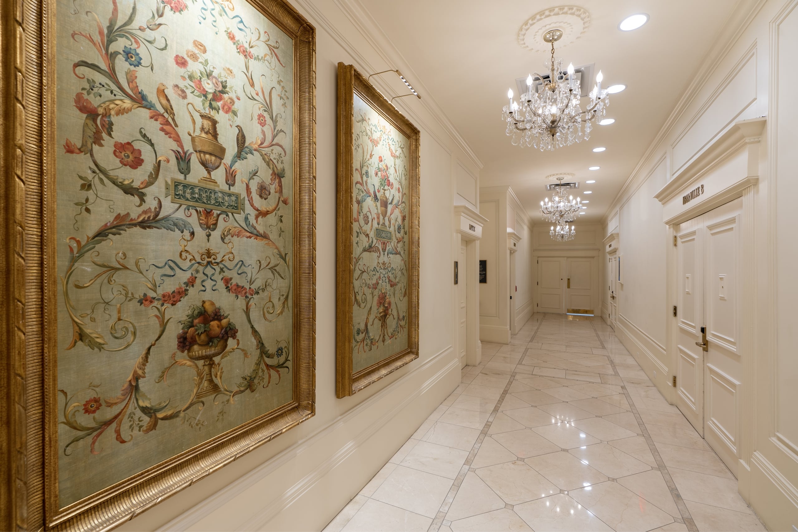 Luxurious hotel hallway with ornate floral paintings in gilded frames, crystal chandeliers, and polished marble floors.