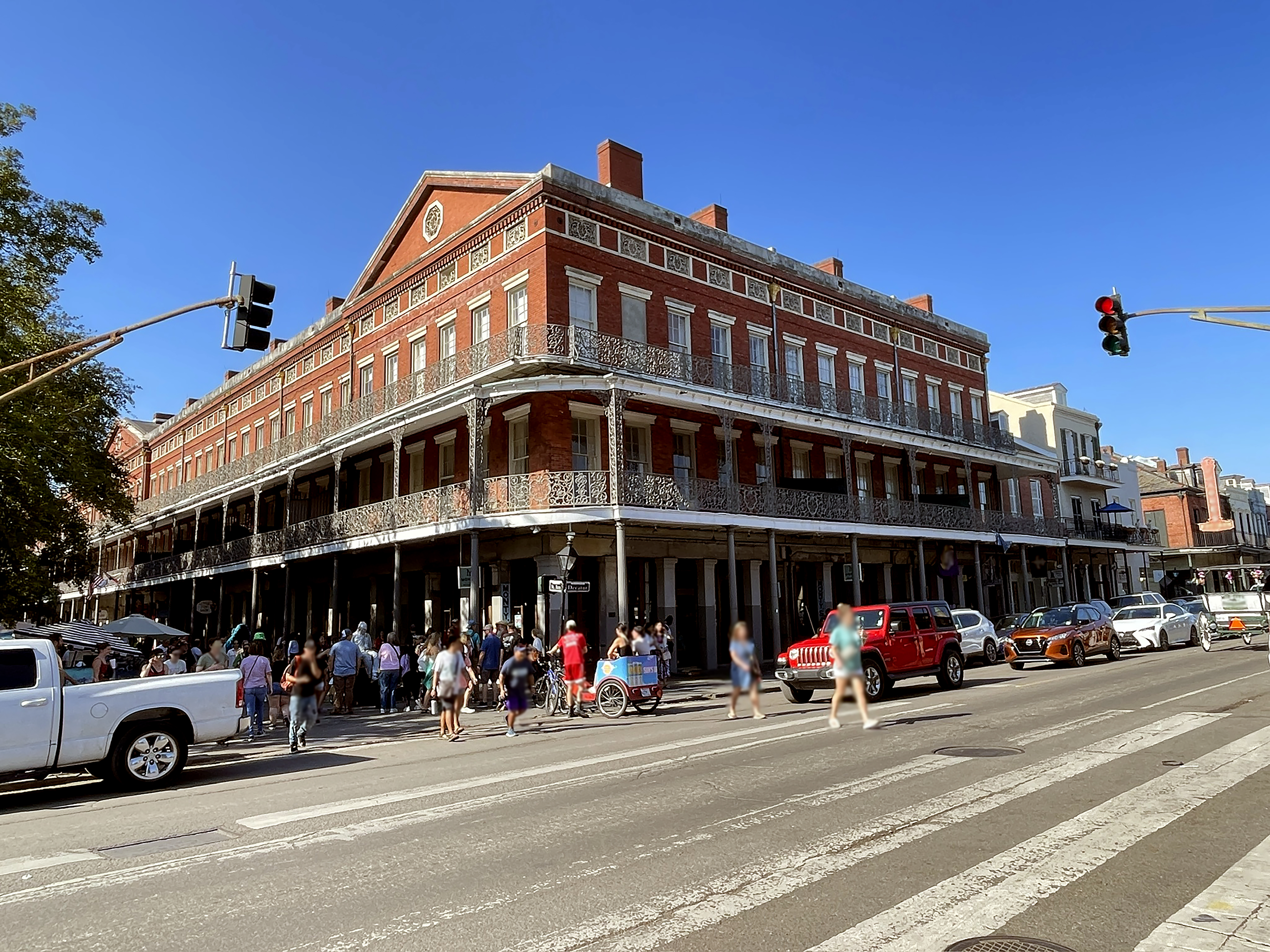 A wide exterior view of a hotel with a red brick facade, ornate balcony, and columns, set against a busy street with people and cars.