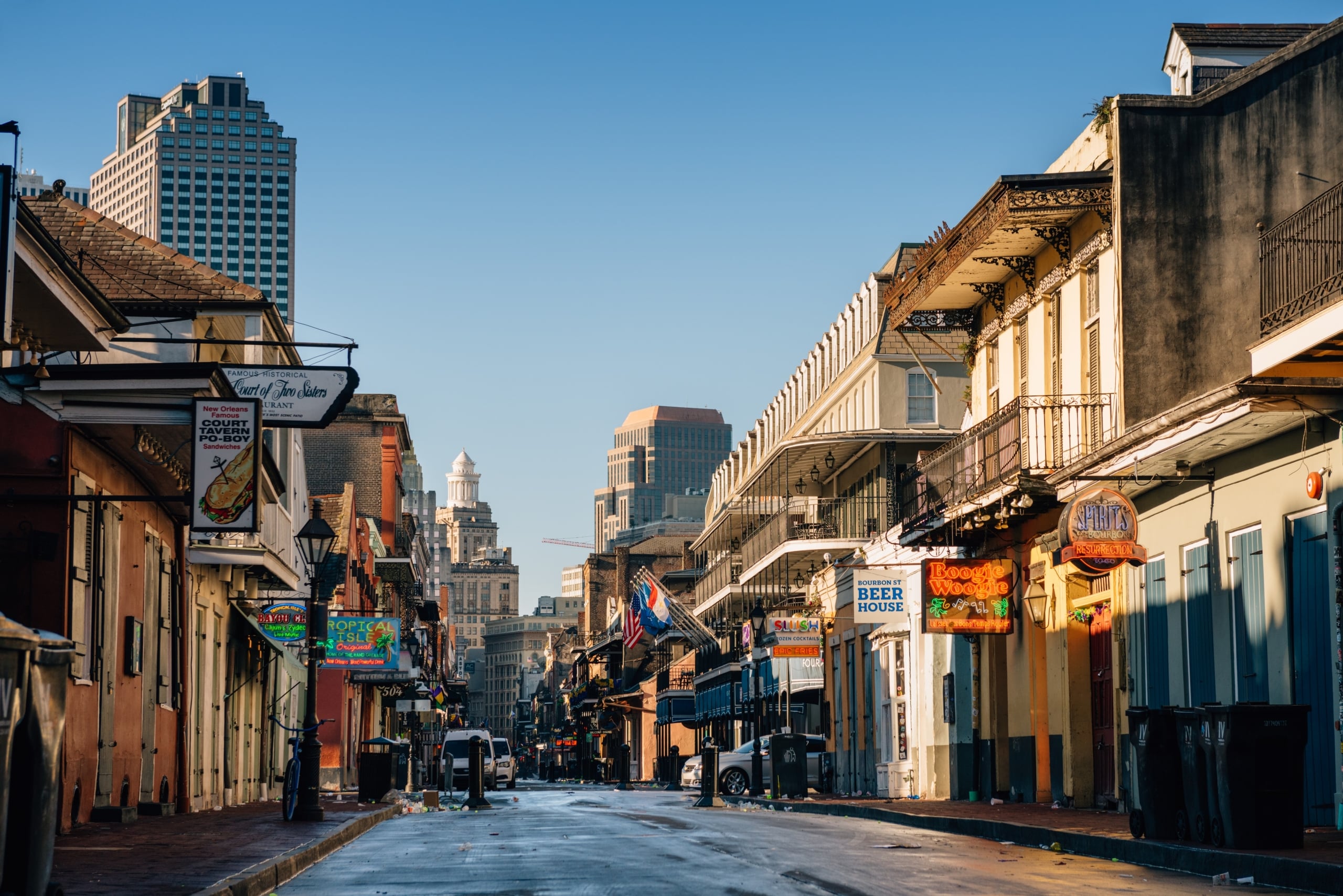 New Orleans' iconic Bourbon Street in the French Quarter, with historic buildings, iron balconies, and vibrant bar signs on a sunny morning.