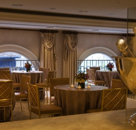 A dining area with round tables, gold chairs, and brown tablecloths, featuring flower arrangements and candles.