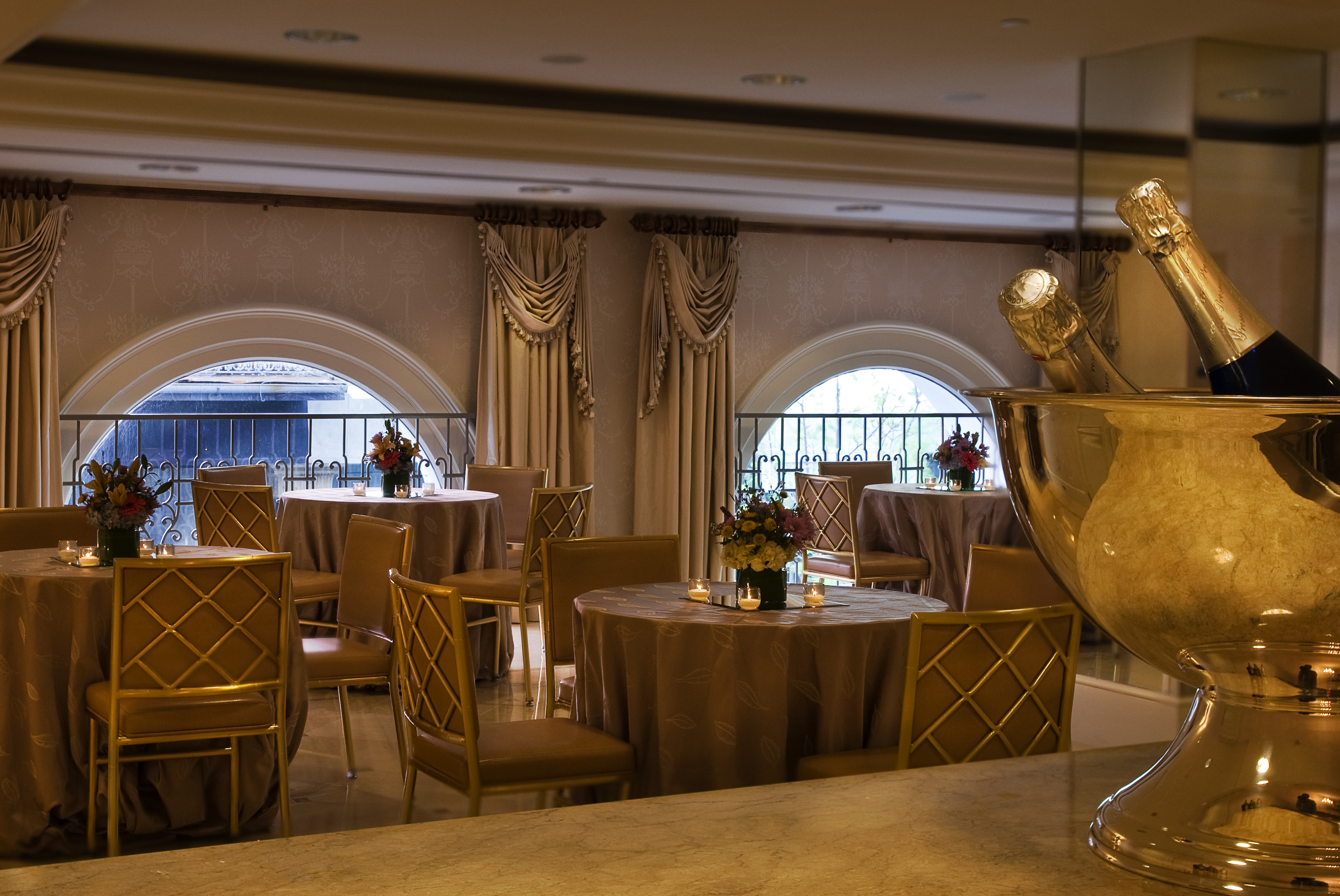 A dining area with round tables, gold chairs, and brown tablecloths, featuring flower arrangements and candles.