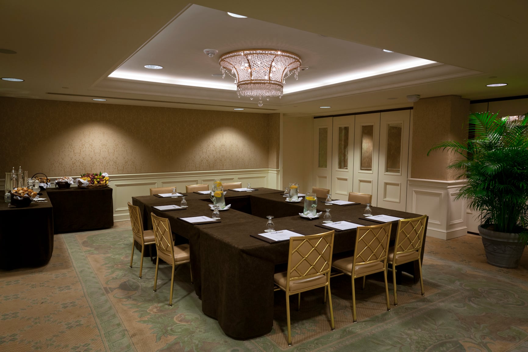 Luxurious conference room with U-shaped table, gold chairs, crystal chandelier, and refreshment station.