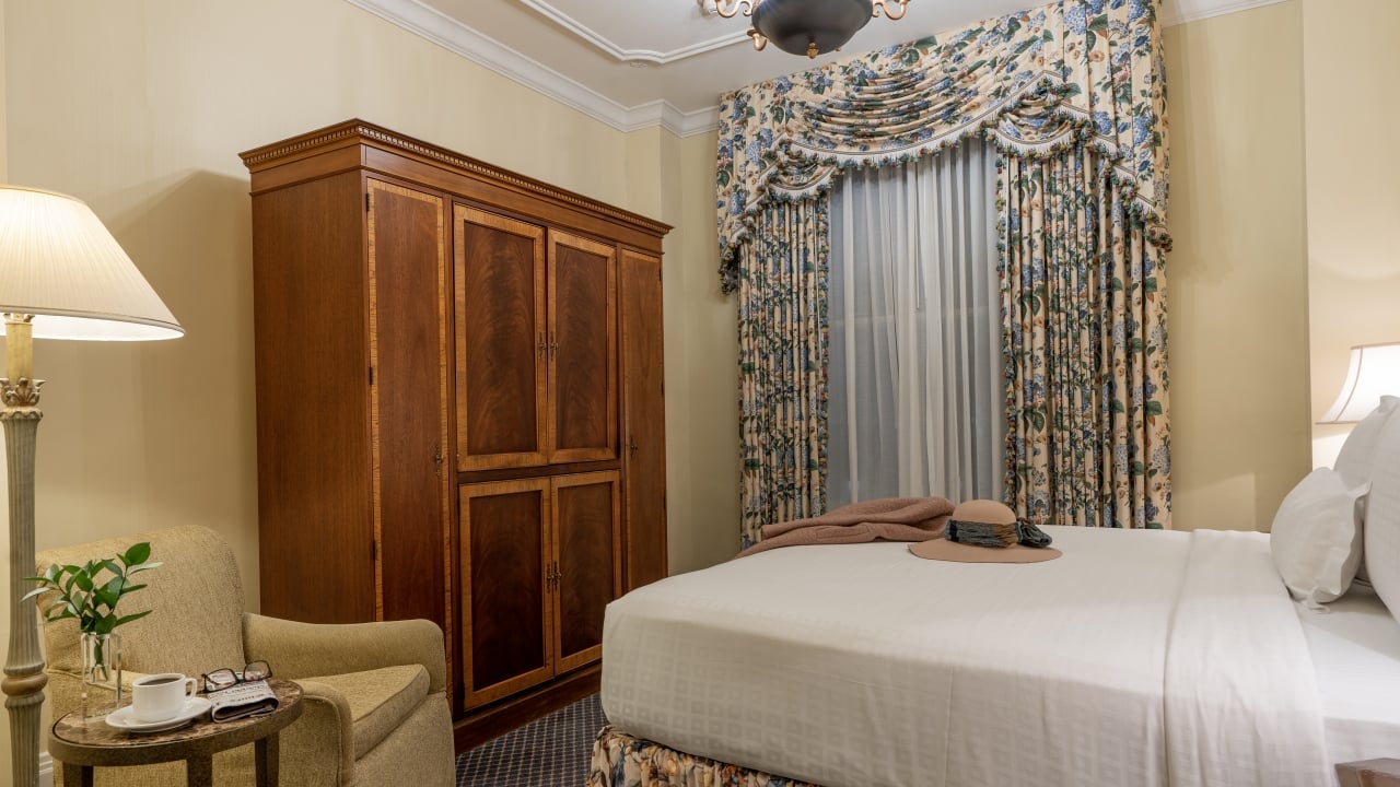 A hotel room with a bed, chair, lamp, wardrobe, and window.