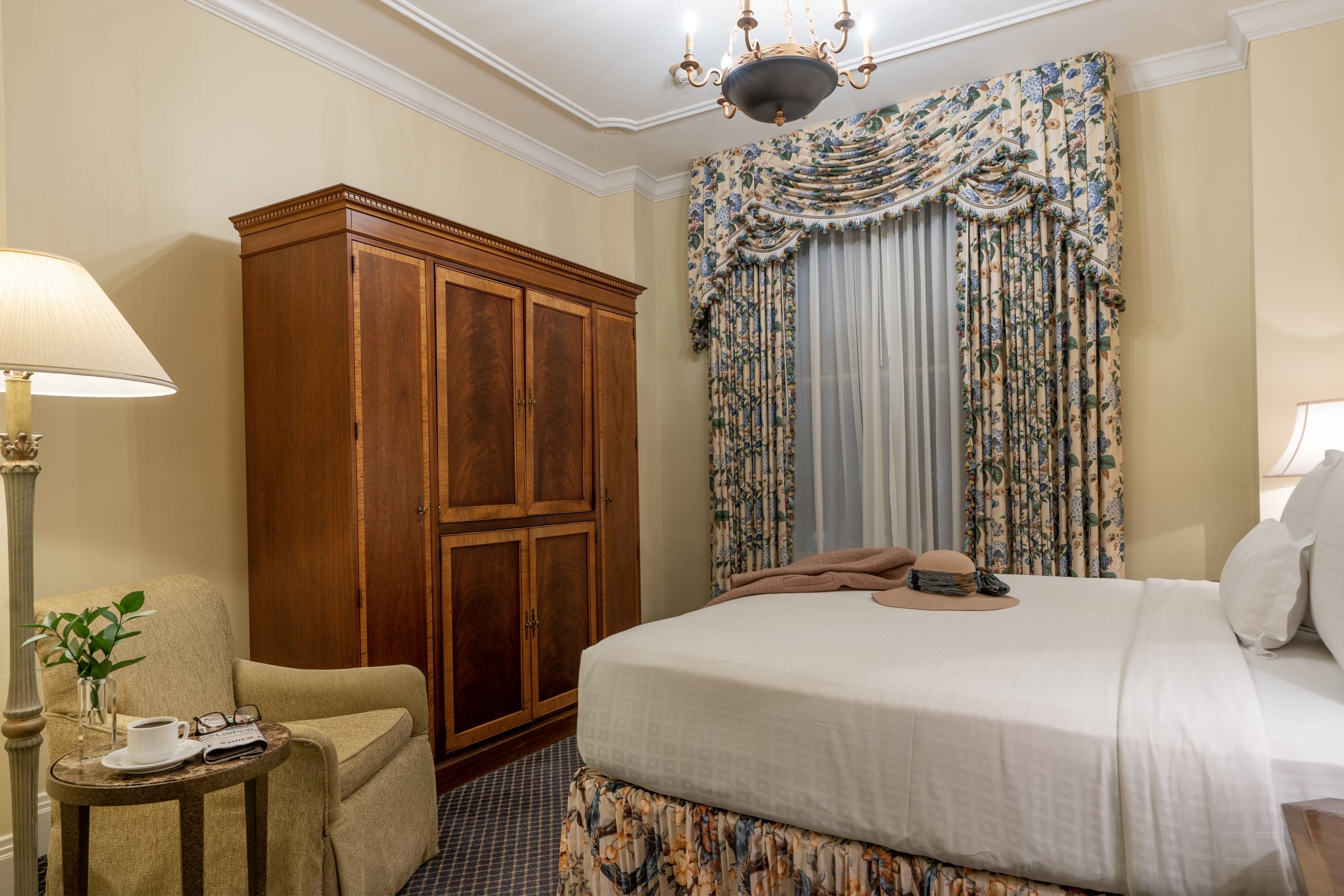 Elegant New Orleans hotel room with a comfortable bed, classic wooden armoire, armchair, and ornate floral curtains.
