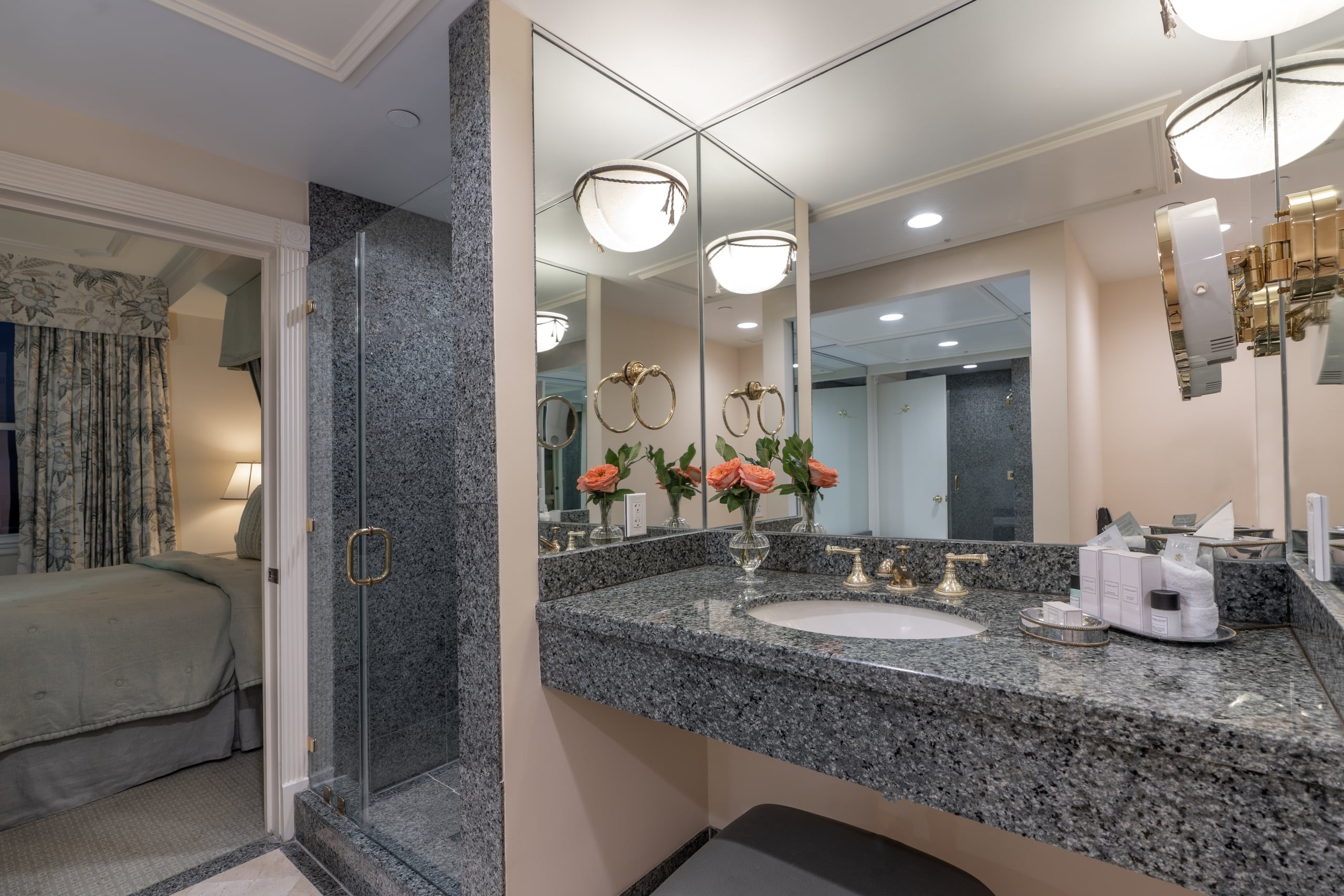Luxurious hotel bathroom with granite vanity, gold fixtures, glass shower, and view into a comfortable bedroom.