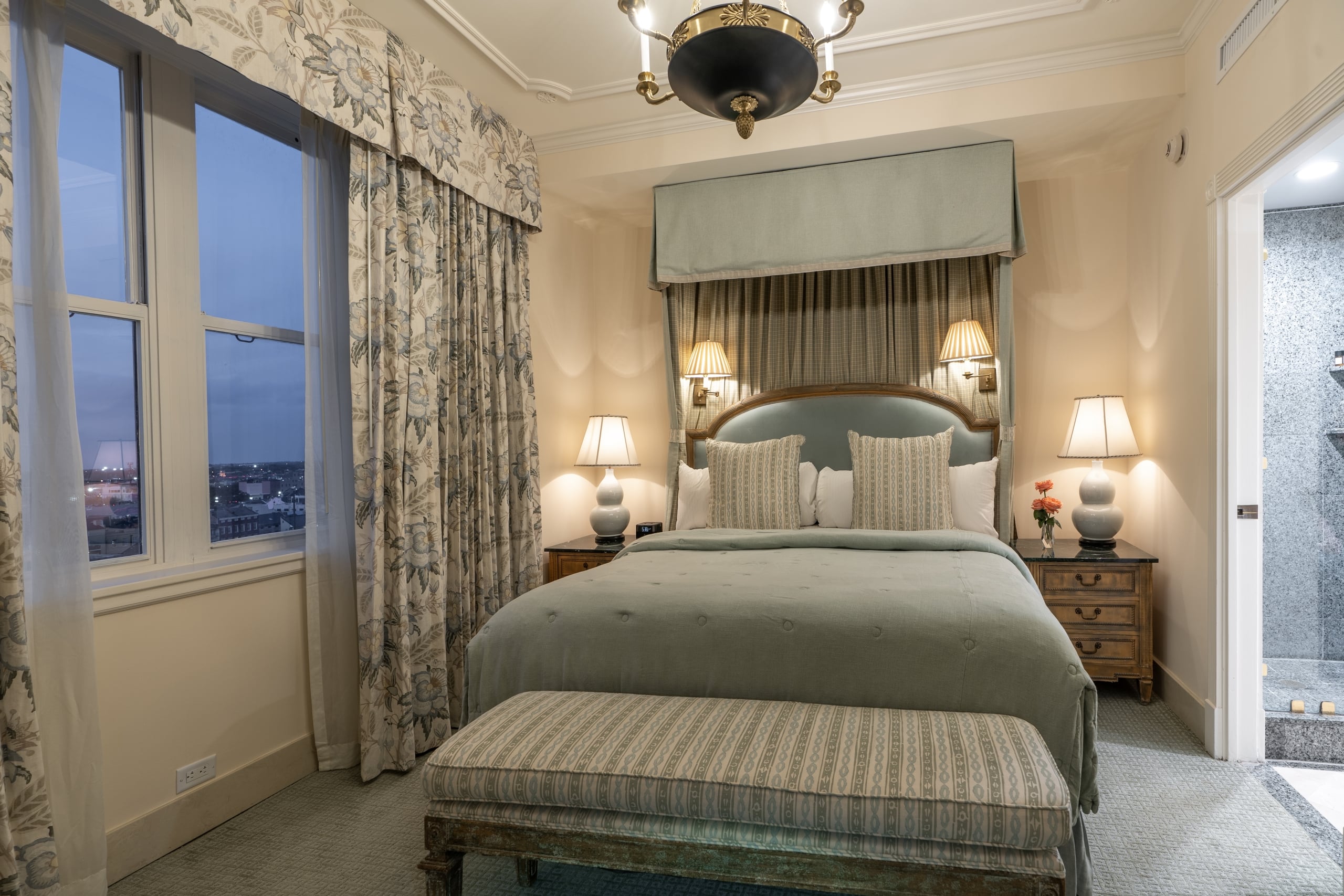 New Orleans luxury hotel bedroom suite with a king bed, ornate headboard, elegant decor, and a scenic city view.