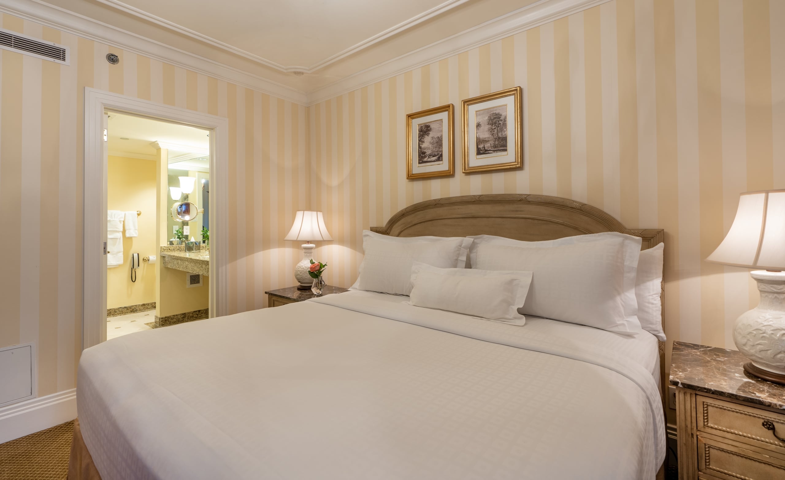 Elegant hotel bedroom with a comfortable bed, classic furniture, striped walls, and a view of the en-suite bathroom.