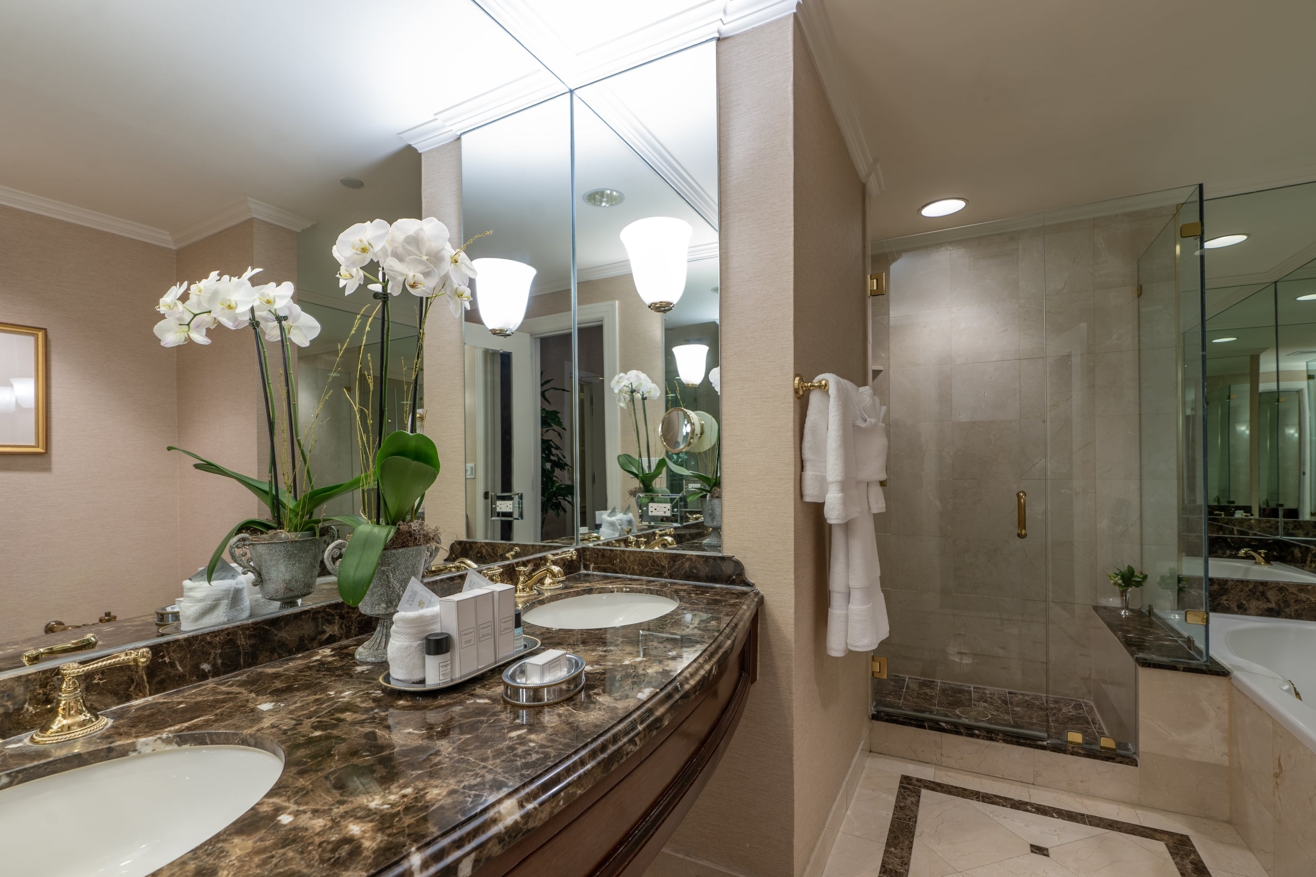 New Orleans luxury hotel bathroom with double marble vanity, gold fixtures, orchids, and glass shower.