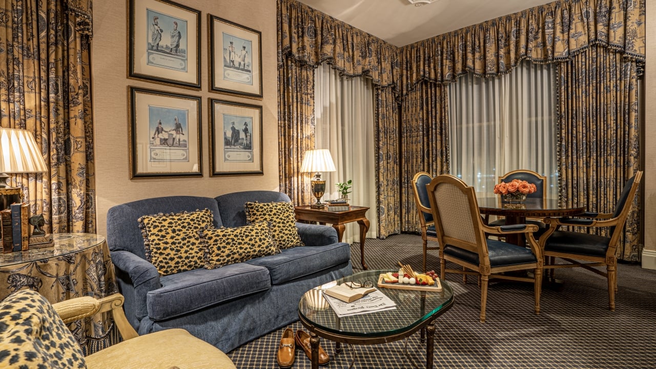 A wide view of a hotel room's living area with a blue sofa, leopard print pillows, a glass coffee table, a dining table with chairs, and framed pictures on the wall.