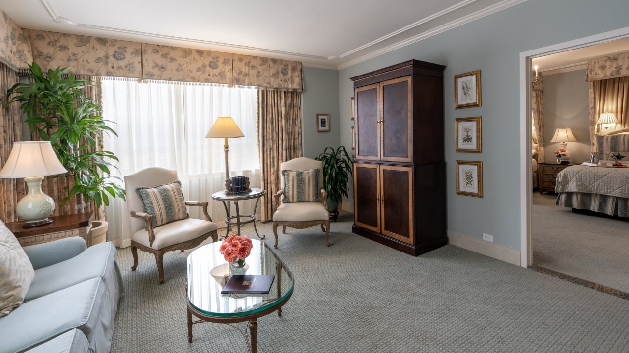 A wide view of a hotel room with a sofa, coffee table, armchairs, and a doorway to the bedroom.