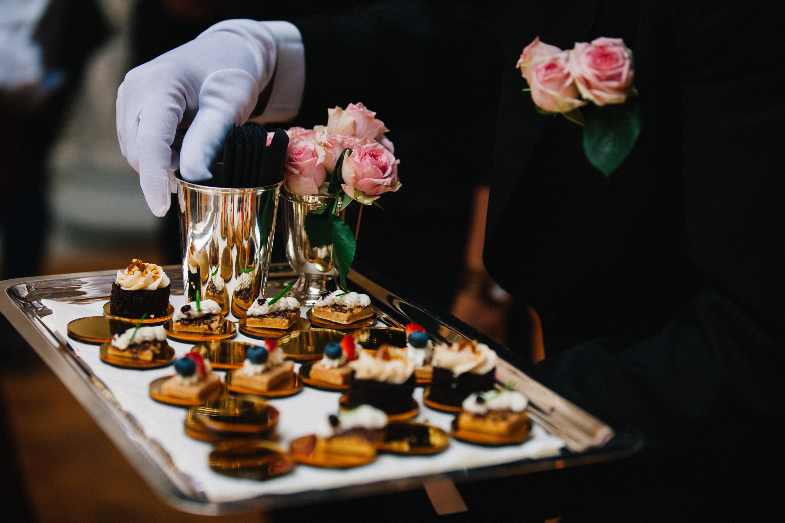 Gloved attendant offers a silver tray with assorted mini desserts and fresh pink roses for New Orleans events.