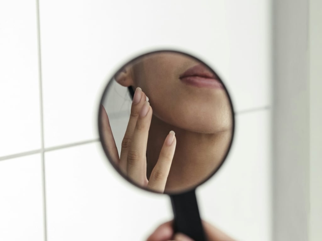 A woman's hand holding a small round mirror, reflecting her face and applying skincare.