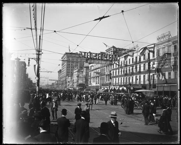 View shows a large number of people gathered on Canal Street, viewed from the intersection of Saint Charles Avenue and Canal Street in the Central Business District. The crowd is gathered in celebration of Armistice