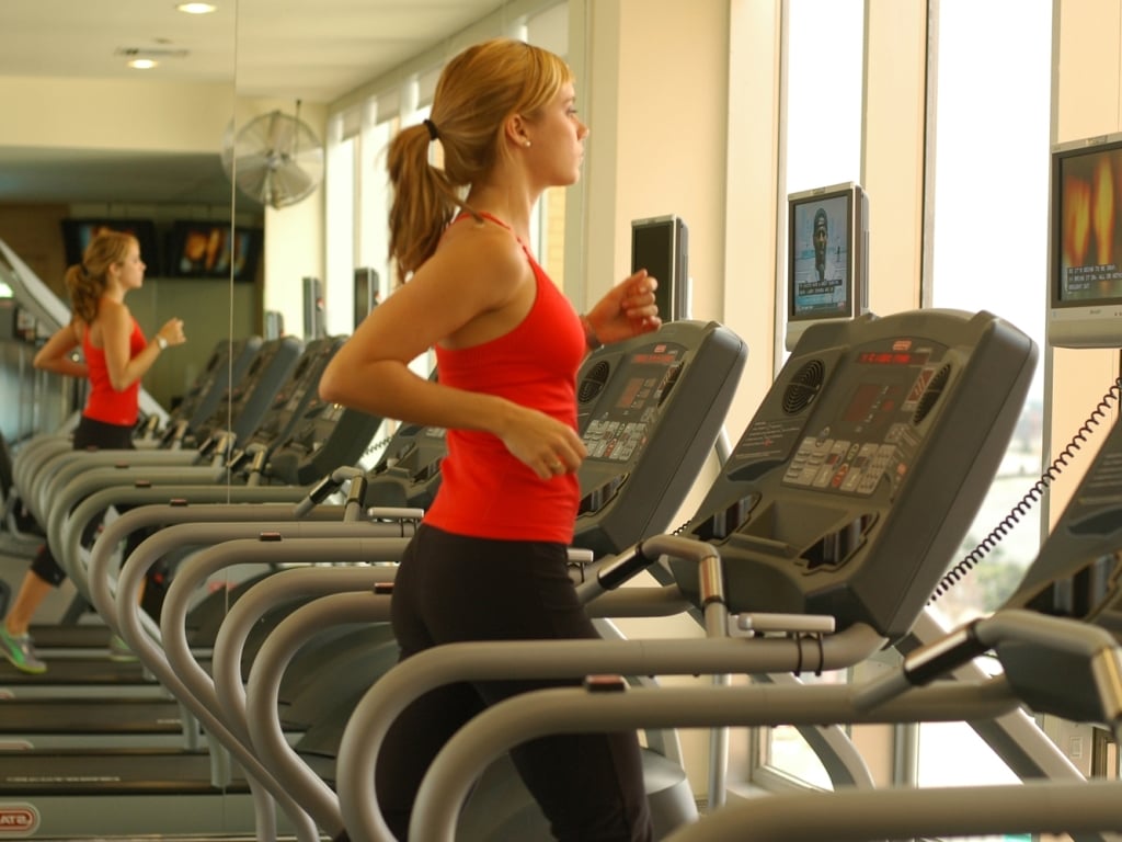 A woman in a red tank top and black leggings runs on a treadmill in a gym with windows and mirrors.