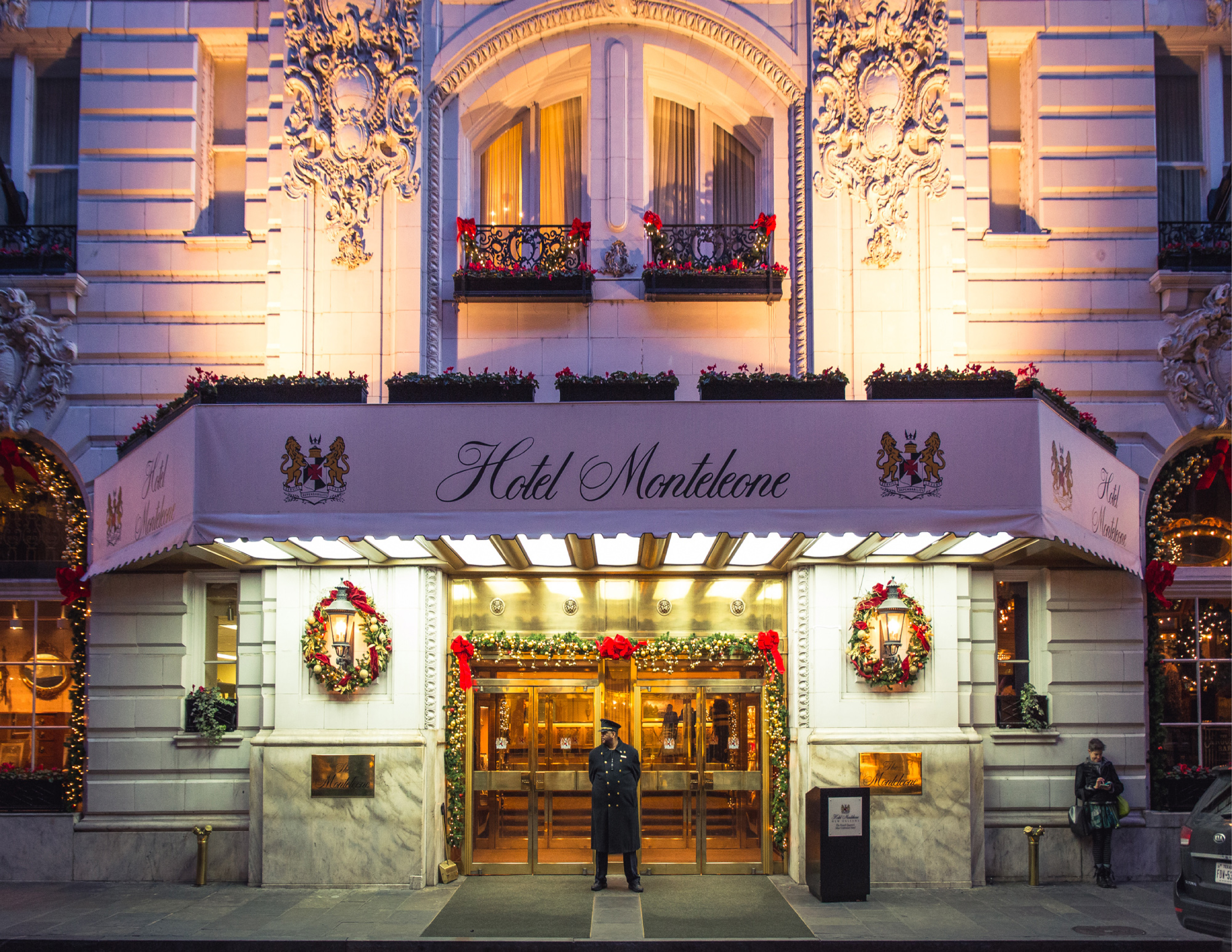 A wide shot of the Hotel Monteleone's exterior, showcasing its ornate facade, balcony, and wreaths, with a doorman standing at the entrance.